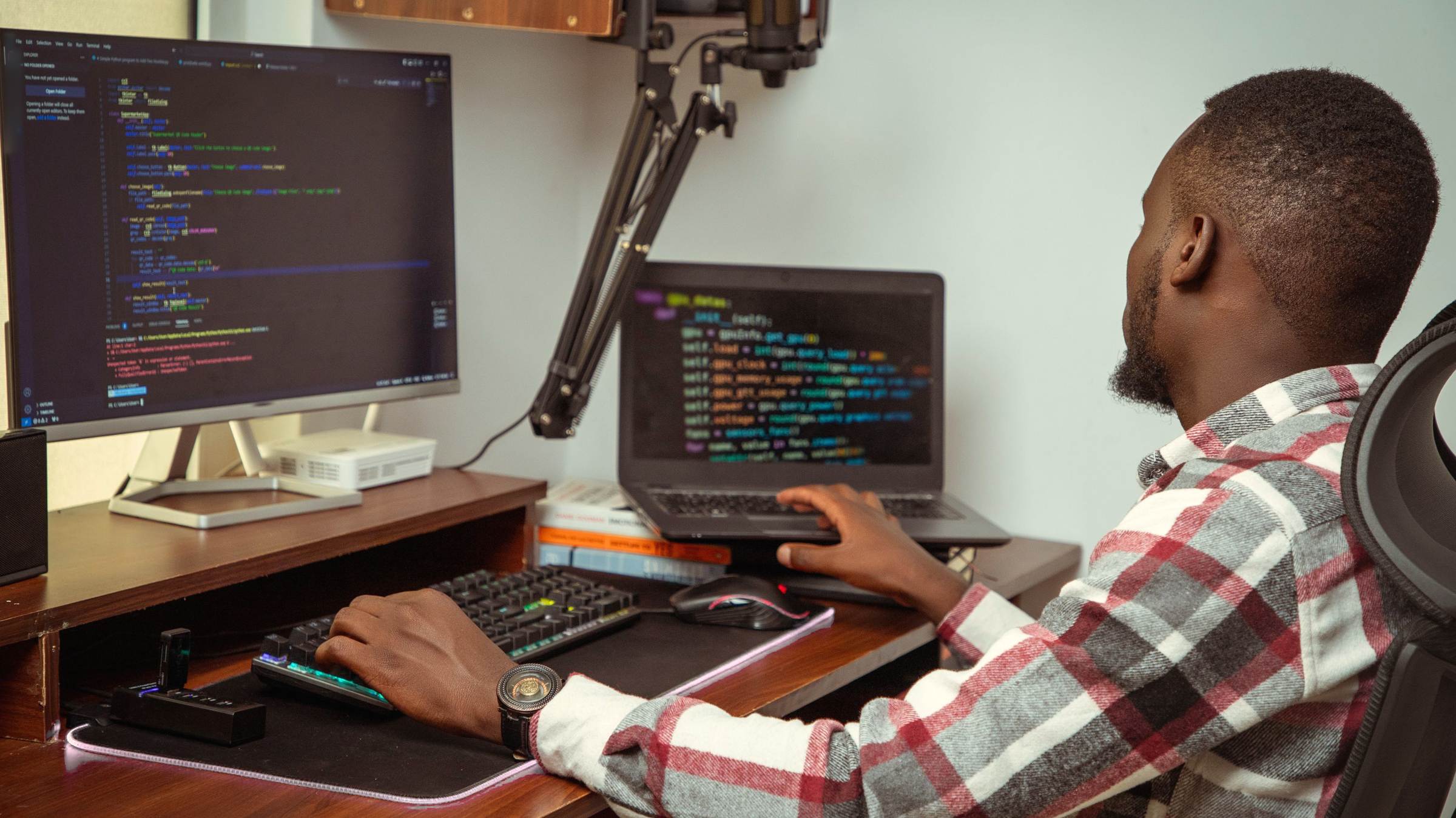 A developer at a desk writes code on a large monitor and a laptop