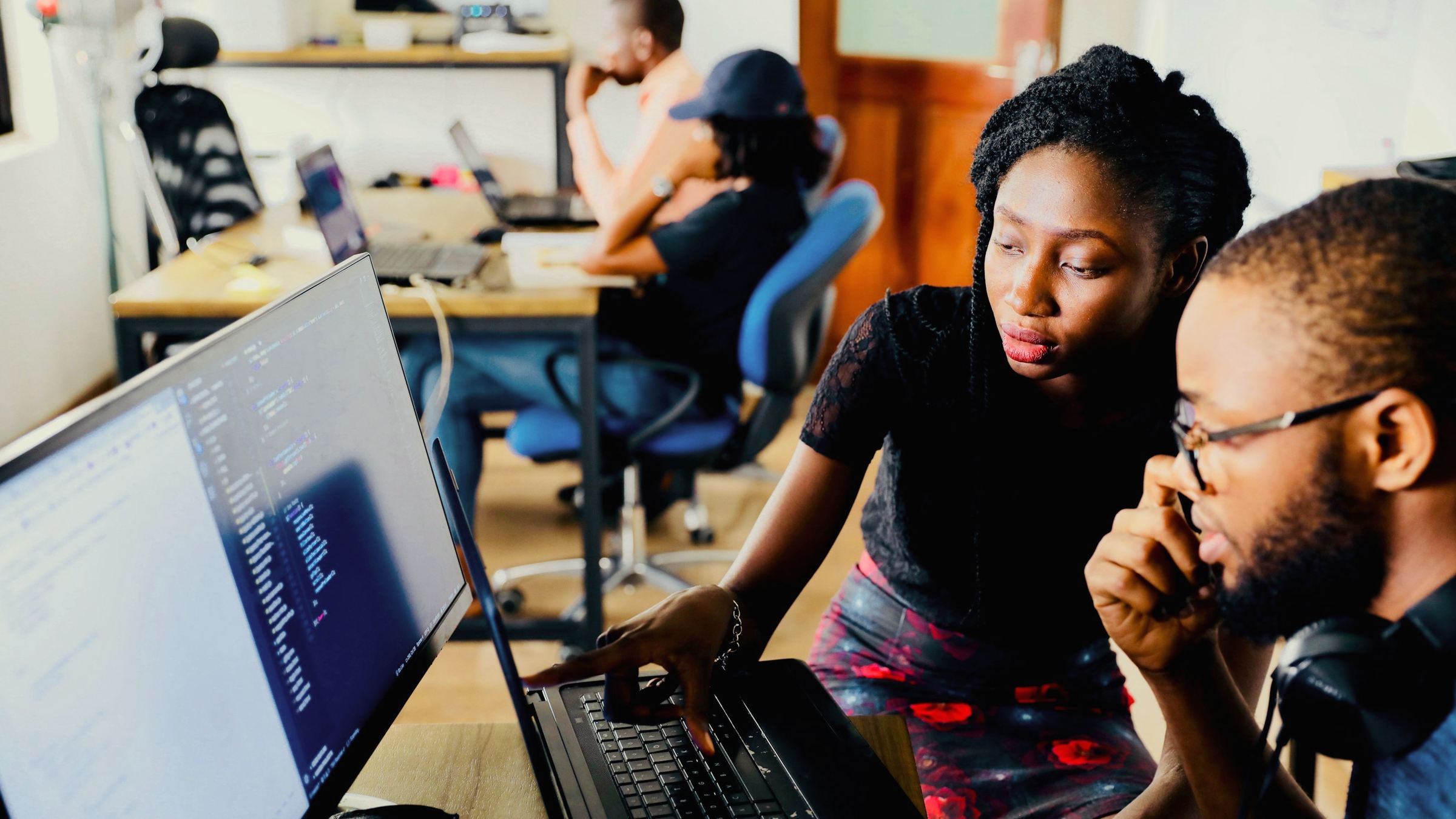 Two developers review code together on a laptop and monitor in a shared office