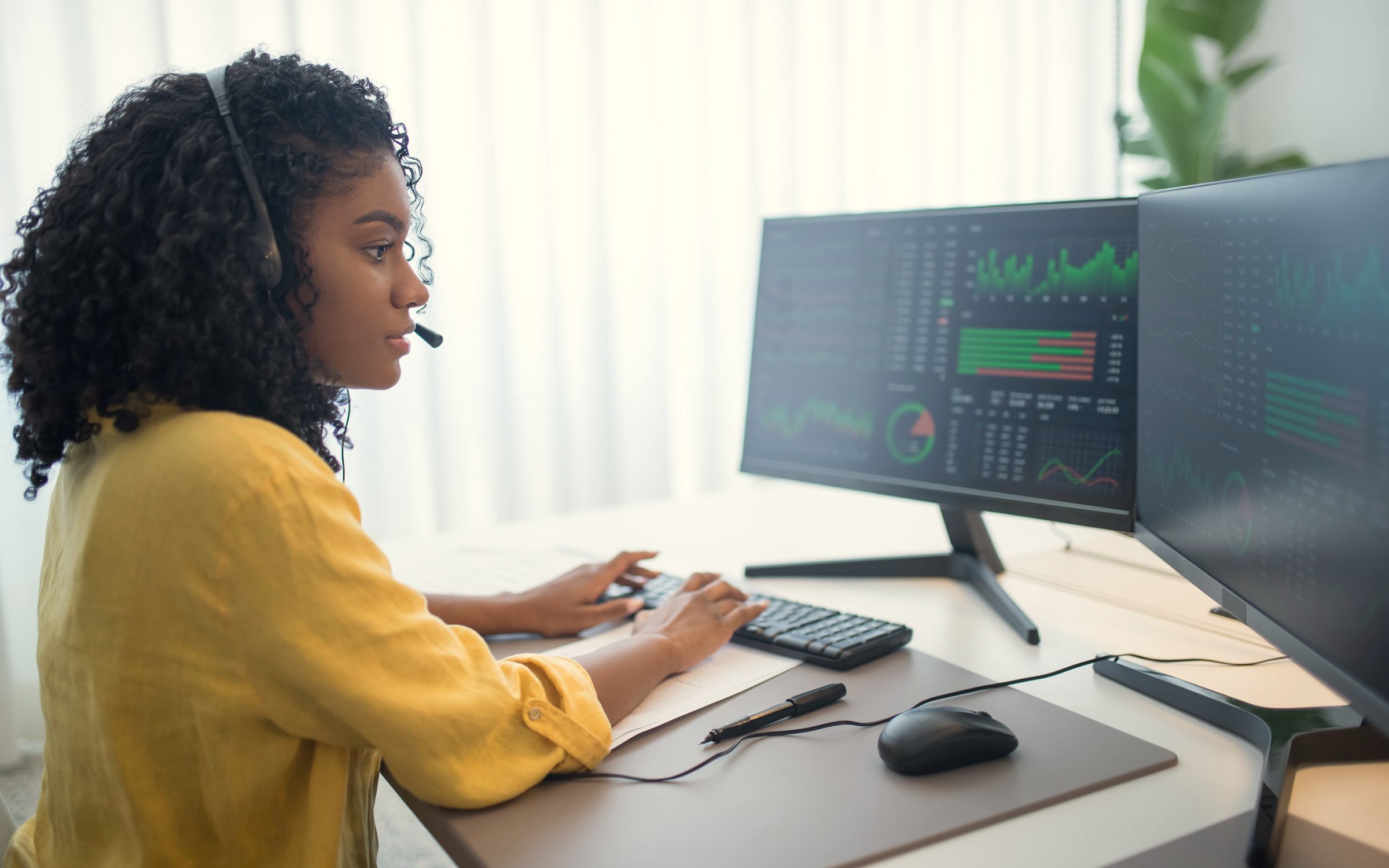 A woman with a headset reviews charts and data tables across two monitors